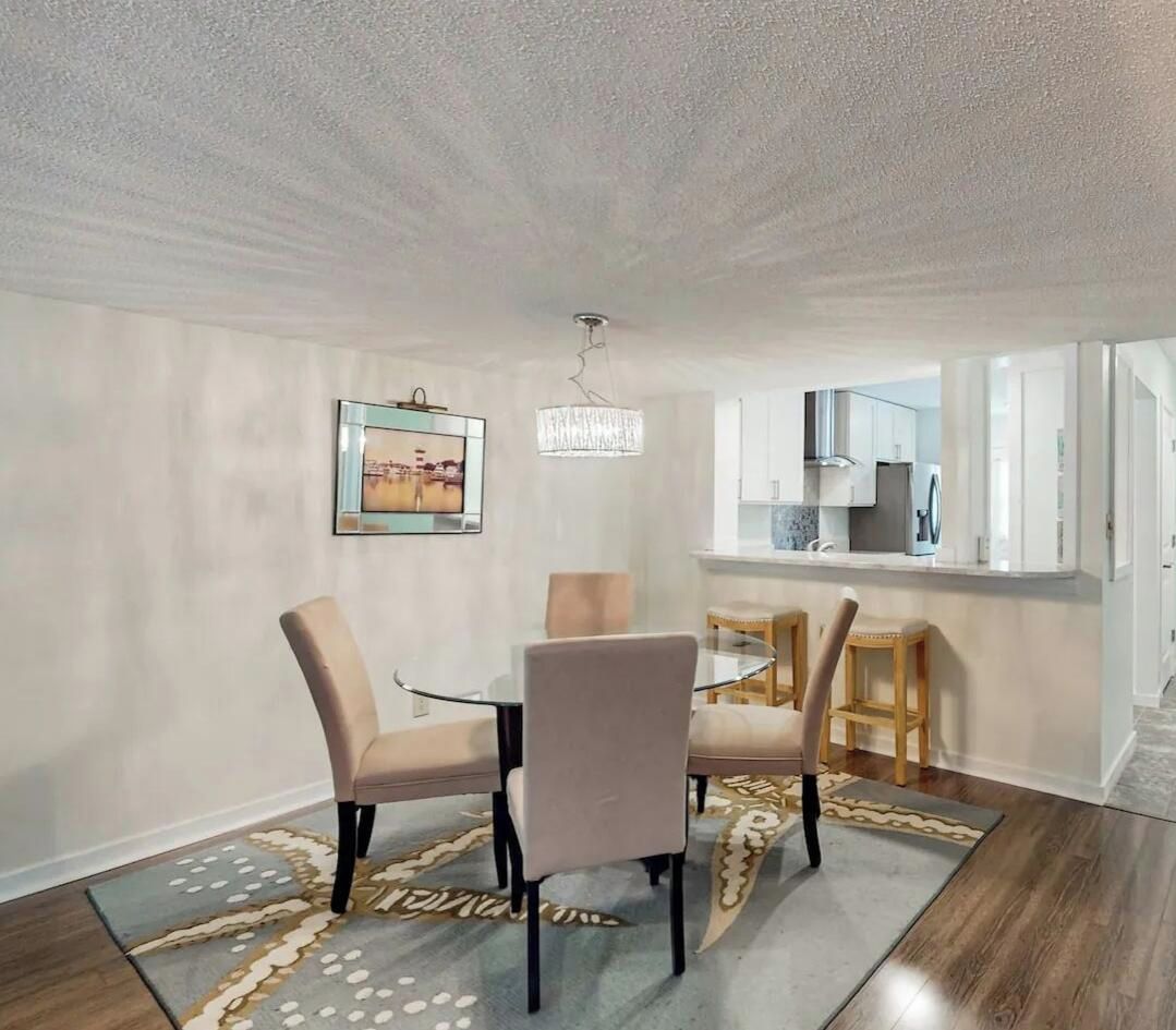 Un coin repas avec une table et des chaises en verre, un lustre, une cuisine en arrière-plan et un tapis à motifs sur le sol.