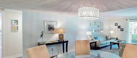 A modern living room with a glass dining table, beige chairs, a sofa, and a chandelier.