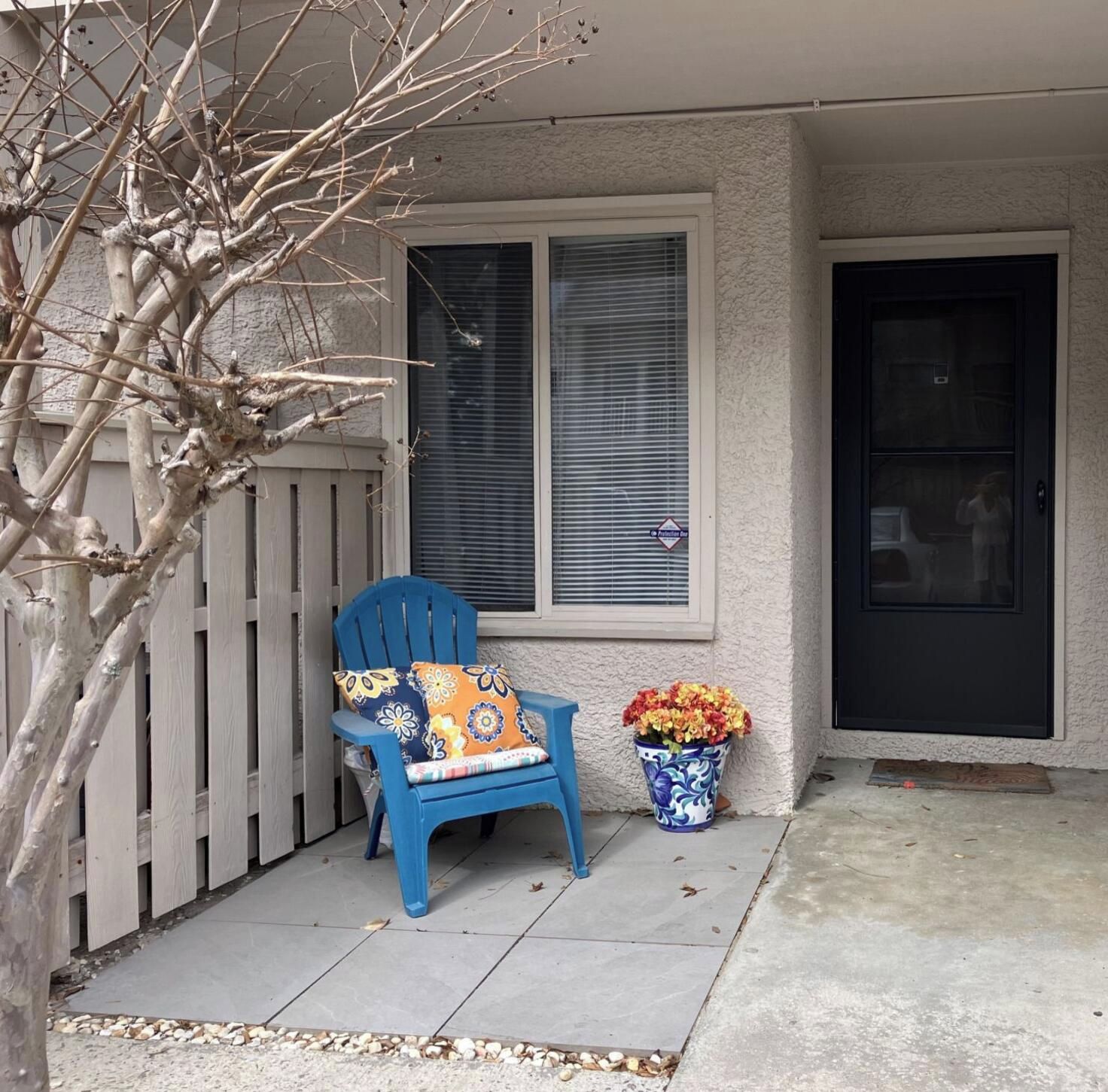A blue chair with a floral cushion outside a house, a potted plant with flowers, and a closed door.