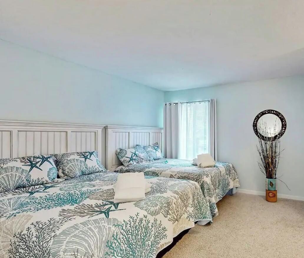 A bedroom with two beds, a window with curtains, a mirror, and a plant.