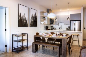 A modern kitchen with a dining area, including a wooden table set for four, a bar stool, and a small bar cart. - Monashee Sunshine (Revelstoke)
