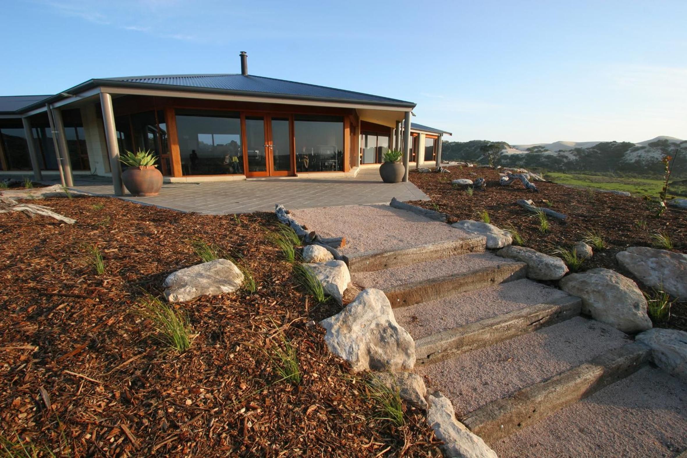 A modern house with a stone staircase leading to a wooden deck, surrounded by a landscaped garden with large rocks and mulch.