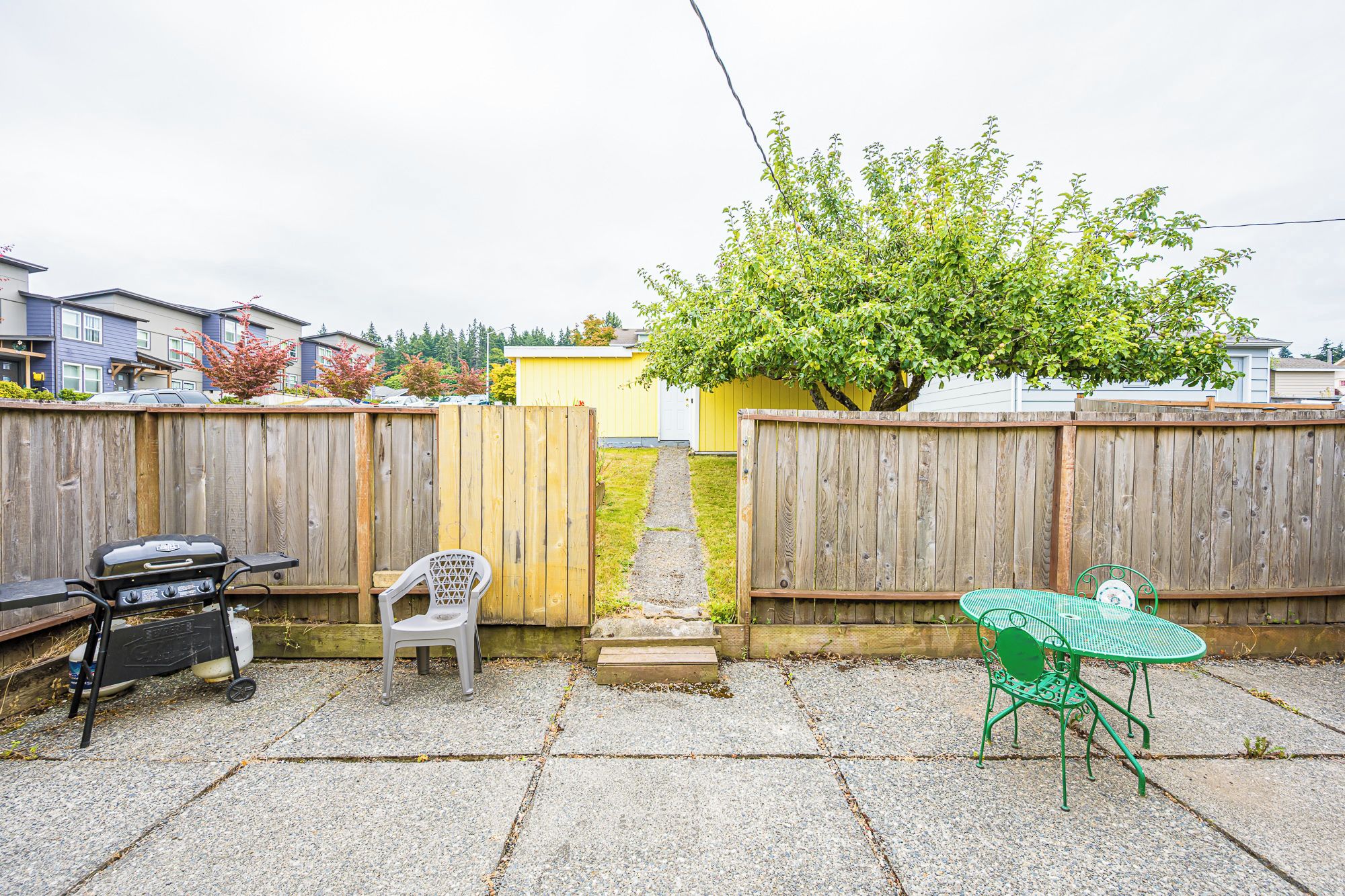 A backyard with a barbecue grill, a green metal table and chair, a white plastic chair, and a wooden fence.