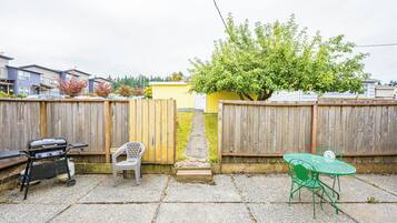 A backyard with a barbecue grill, a green metal table and chair, a white plastic chair, and a wooden fence.