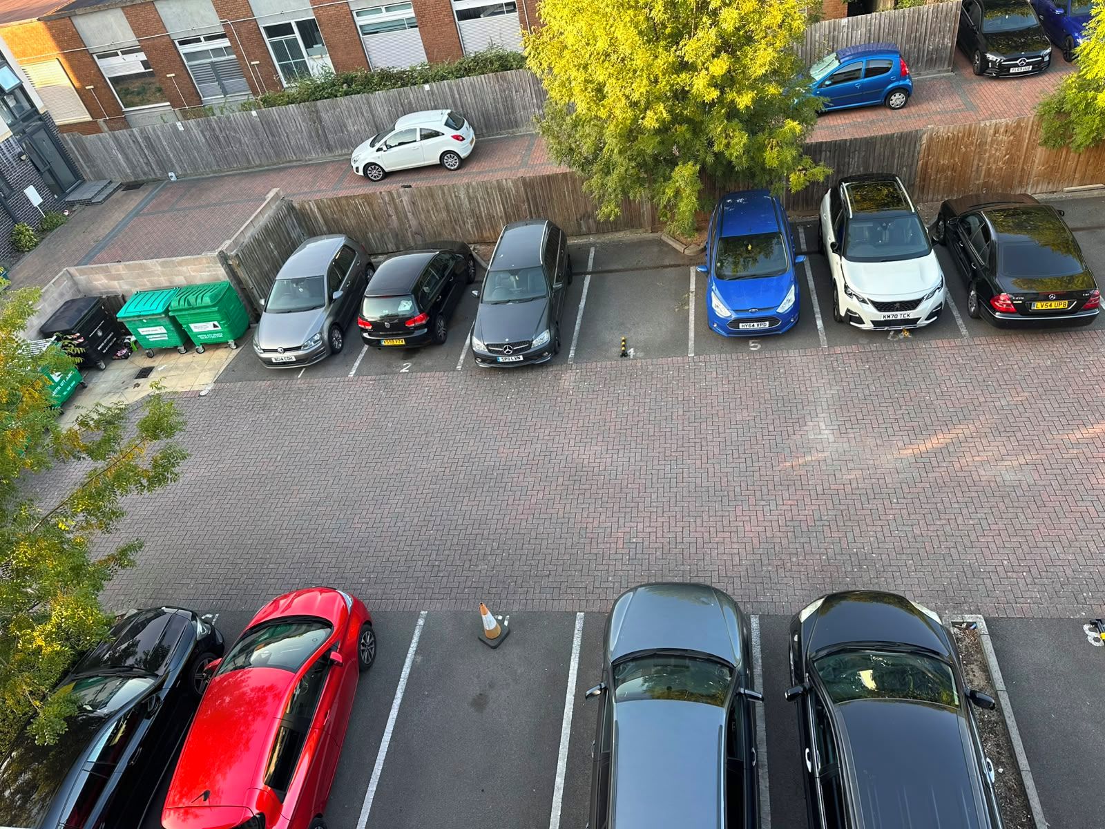 Un estacionamiento con varios autos y contenedores verdes para reciclaje.