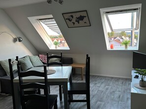 Ein Zimmer mit Esstisch, Stühlen, einer Bank, einer Couch, zwei Oberlichtern, einer Weltkarte und einem Fernseher.