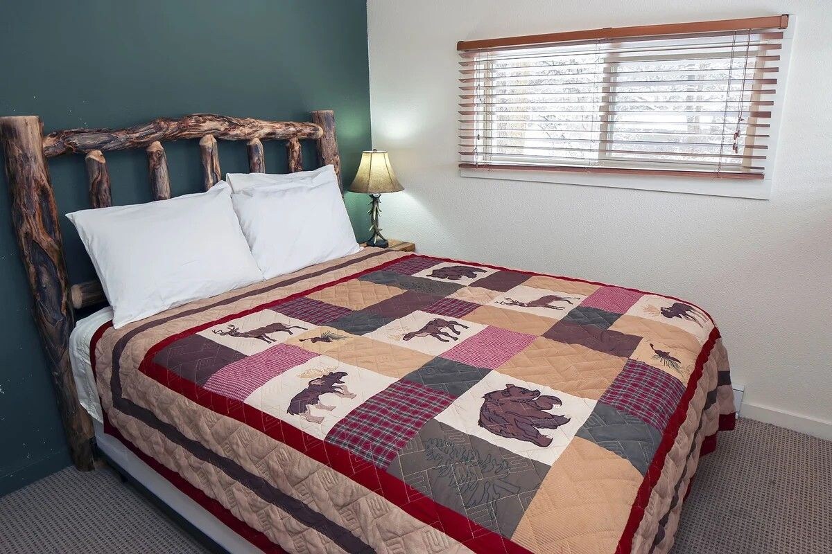 A bed with a wooden headboard, a quilt with animal prints, a bedside lamp, and a window with blinds.
