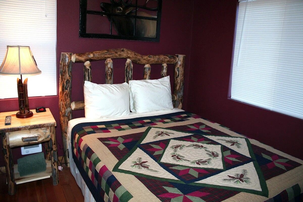 A bedroom with a wooden headboard, a bed with white pillows, a quilt with geometric patterns, a bedside lamp, and a small table with a remote control.
