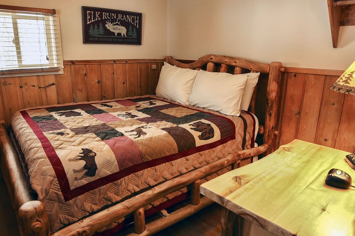 A wooden bed with a quilt featuring elk and deer, a wooden headboard, and a small table with a lamp and mouse.
