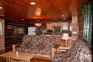 A log cabin interior with a wooden ceiling, a patterned sofa, a small table, and a kitchen area with wooden cabinets. - Triple R Cottages (Estes Park)