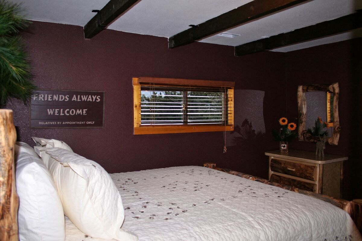 A bedroom with a bed, a window with blinds, a sign on the wall, and a dresser with a mirror and flowers.