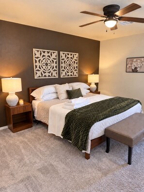 Quarto com cama, luminárias de cabeceira, painéis decorativos na parede, ventilador de teto e piso coberto por carpete.