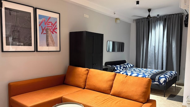 Una habitación de hotel moderna con sofá, una pequeña mesa redonda, una cama con ropa de cama estampada, un televisor de pantalla plana y dos cuadros en la pared.