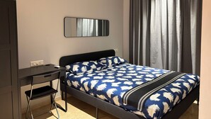 Un dormitorio con cama, escritorio, silla, ventilador de techo, espejo y ventana con cortinas.