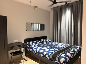 Een slaapkamer met een bed, een bureau, een stoel, een plafondventilator, een spiegel en een raam met gordijnen.