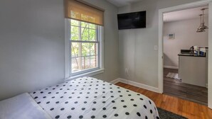 Een slaapkamer met een bed, een raam met jaloezieën, een televisie aan de muur en een kleine kitchenette.