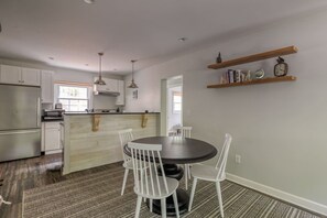 Una cocina moderna con armarios blancos, una isla central, iluminación colgante y una zona de comedor con una mesa redonda y sillas.