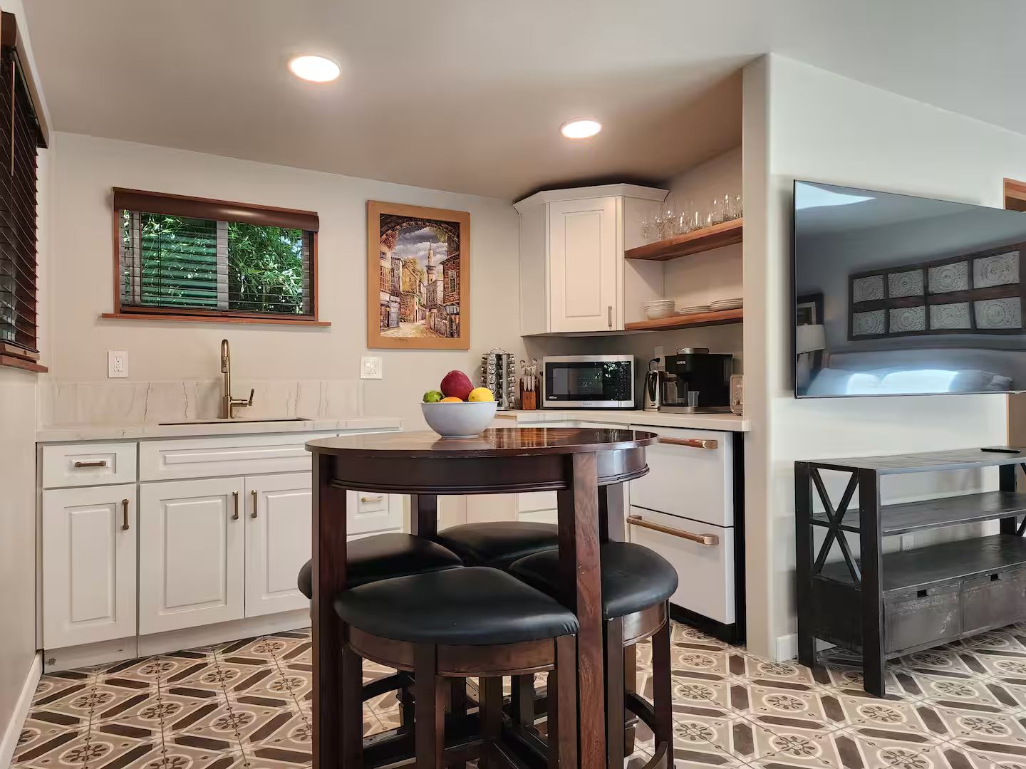 Cozinha moderna com armários brancos, área de jantar com mesa de madeira e cadeiras pretas, micro-ondas e televisão na parede.