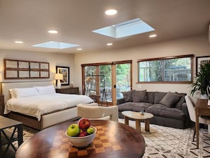 Ein modernes Schlafzimmer mit Bett, Nachttisch und Beistelltisch. Es gibt ein Sofa, einen kleinen runden Tisch mit einer Obstschale und ein Fenster mit Jalousien.