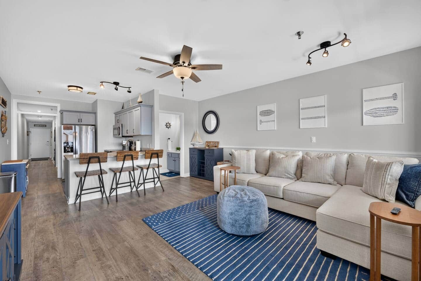 A modern living room with a sofa, ottoman, and a coffee table. There is a kitchen area with a refrigerator and bar stools.