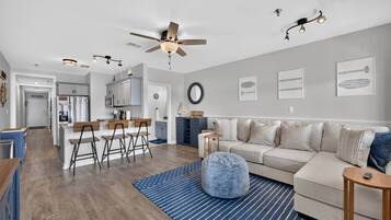 A modern living room with a sofa, ottoman, and a coffee table. There is a kitchen area with a refrigerator and bar stools.