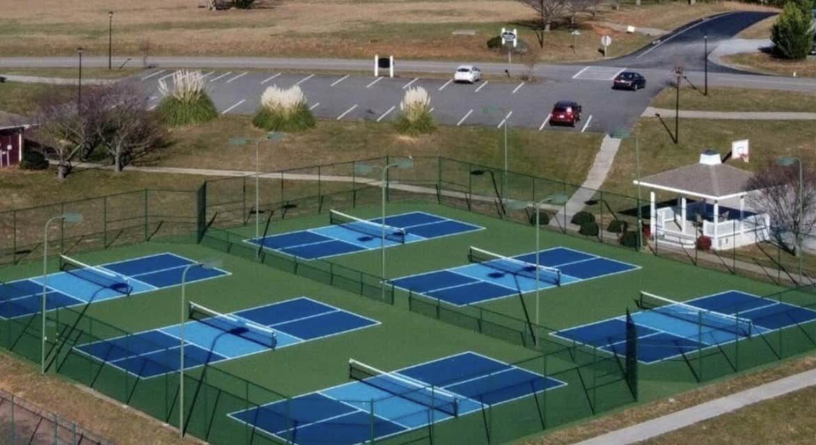 Un complexe de courts de tennis en plein air comprenant plusieurs terrains, un espace de stationnement et un petit bâtiment.