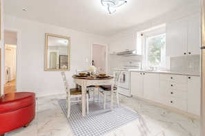 A modern kitchen with white cabinetry, a dining table with chairs, a red ottoman, and a large window providing natural light. - Family 2-bedroom near Ottawa – Wi-Fi and free parking (Gatineau)