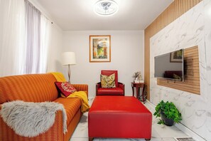 A modern living room with a red sofa, a red ottoman, a red armchair, a floor lamp, a framed picture on the wall, and a TV mounted on the wall. - Family 2-bedroom near Ottawa – Wi-Fi and free parking (Gatineau)
