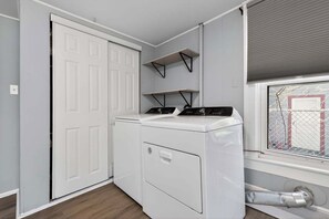 A laundry room with a front-loading washer and dryer, a white door, and a window with blinds. - Cozy Southfield Ranch (Southfield)