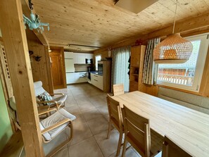 Interior de madeira com mesa de jantar, cadeiras, área de cozinha e janela com cortinas.