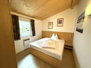 Quarto com cabeceira de madeira, cama com roupa de cama branca, janela com cortinas e duas fotos emolduradas na parede.