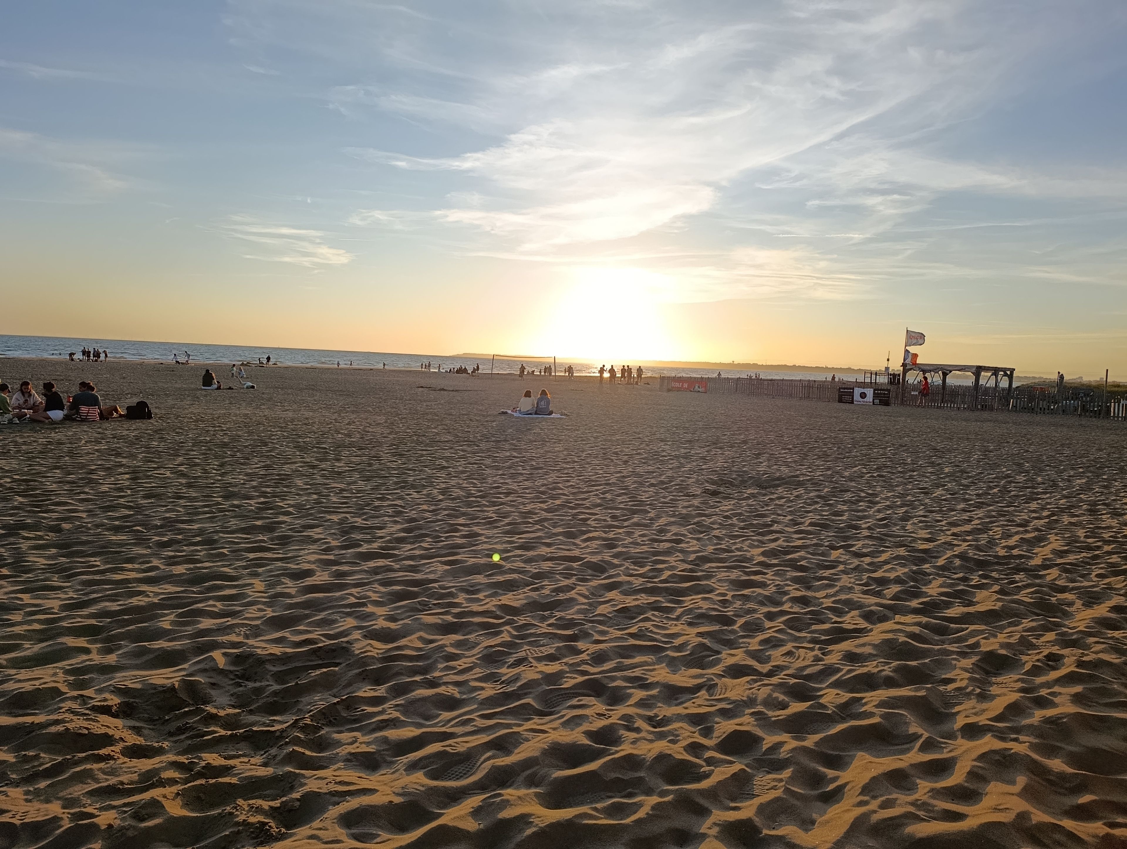 A beach with people sitting and walking, a lifeguard tower, and a wooden fence.