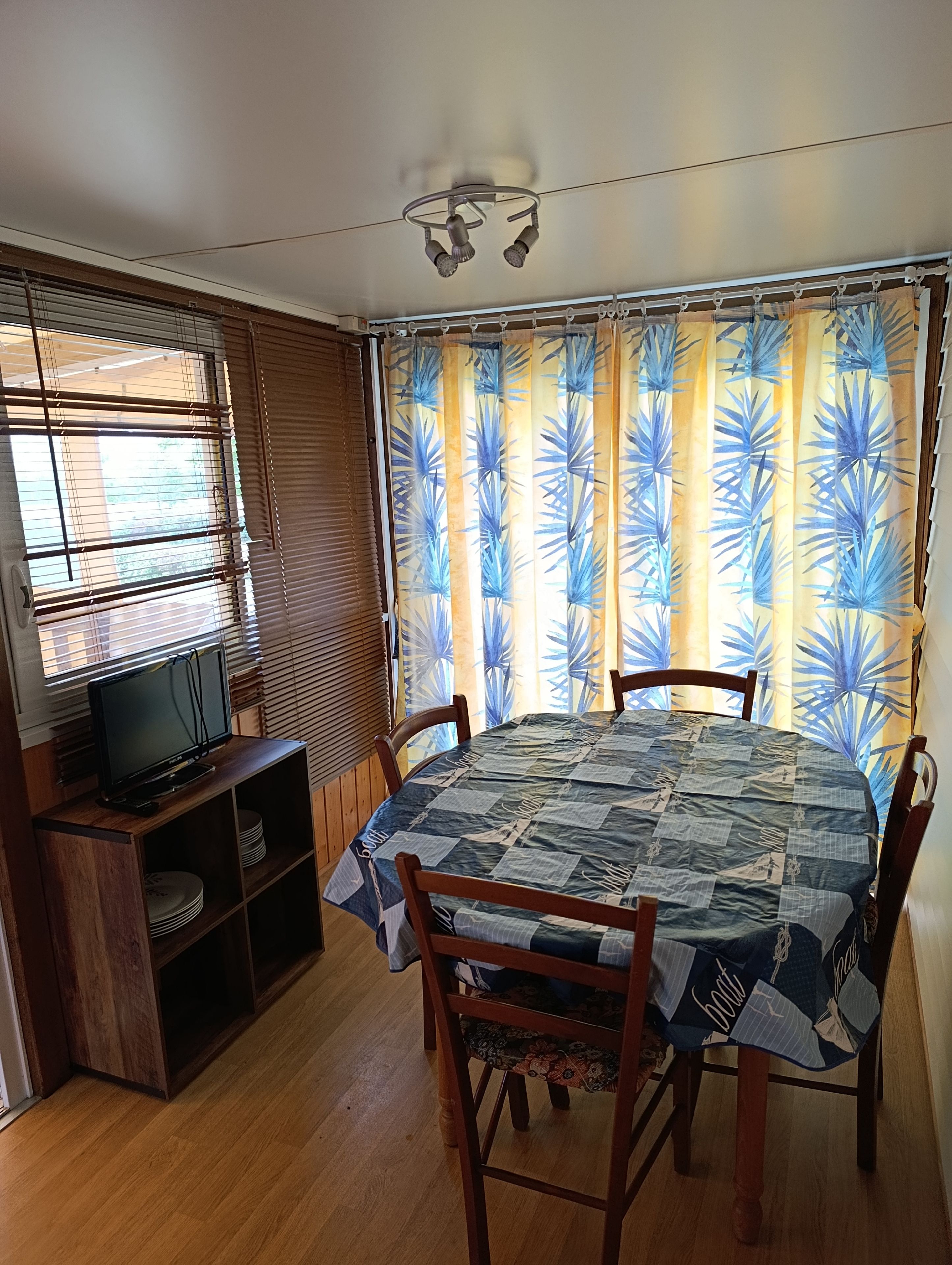A dining area with a round table covered by a checkered tablecloth, flanked by wooden chairs. A television is placed on a wooden stand to the left.