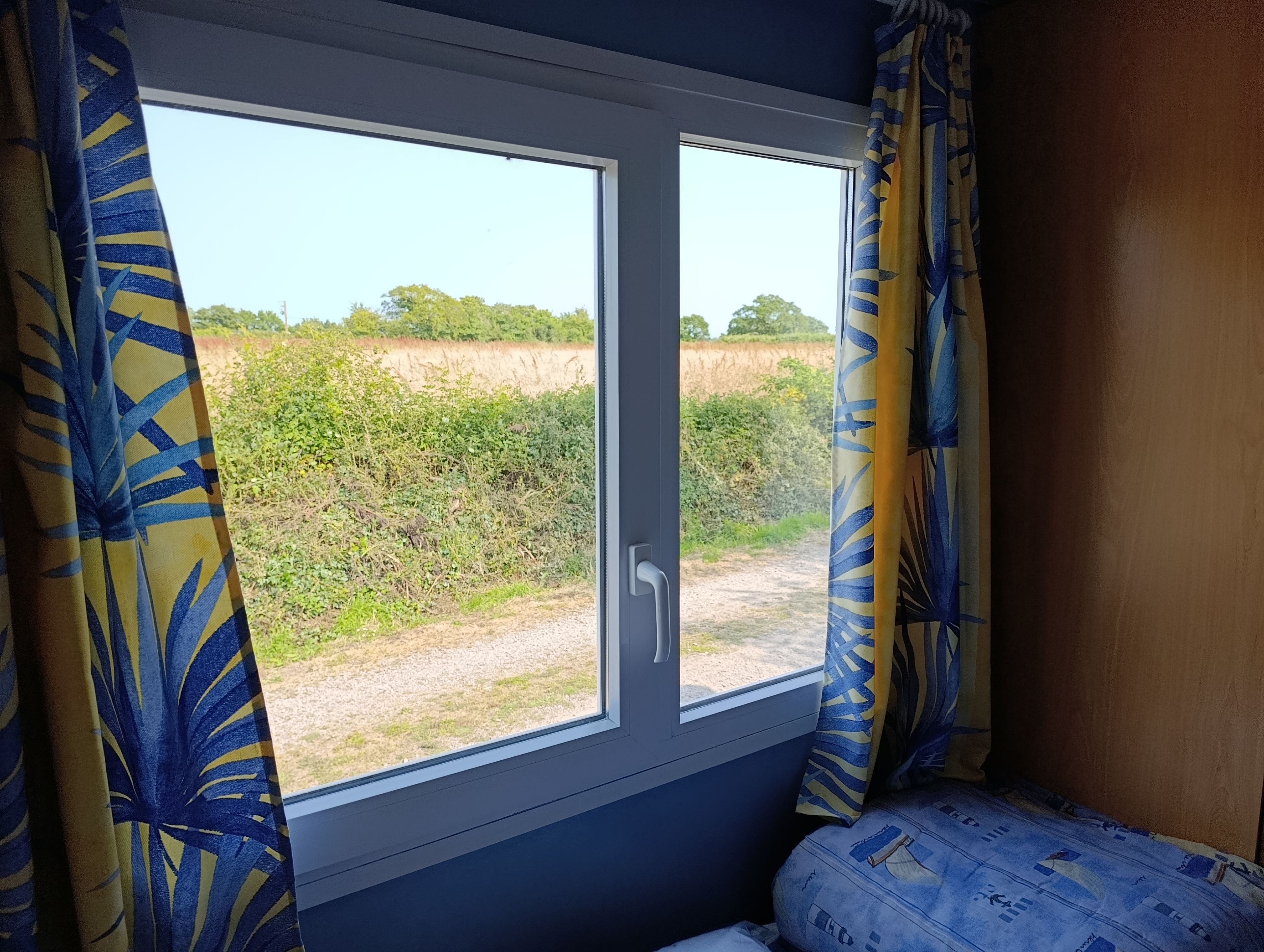 Een kamer met een raam dat uitkijkt op een landelijk landschap, een bed met beddengoed met een patroon en gordijnen met een bladmotief.