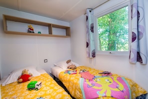 Een slaapkamer met twee bedden, een raam met gordijnen, een plank met speelgoed, en een teddybeer op een van de bedden.