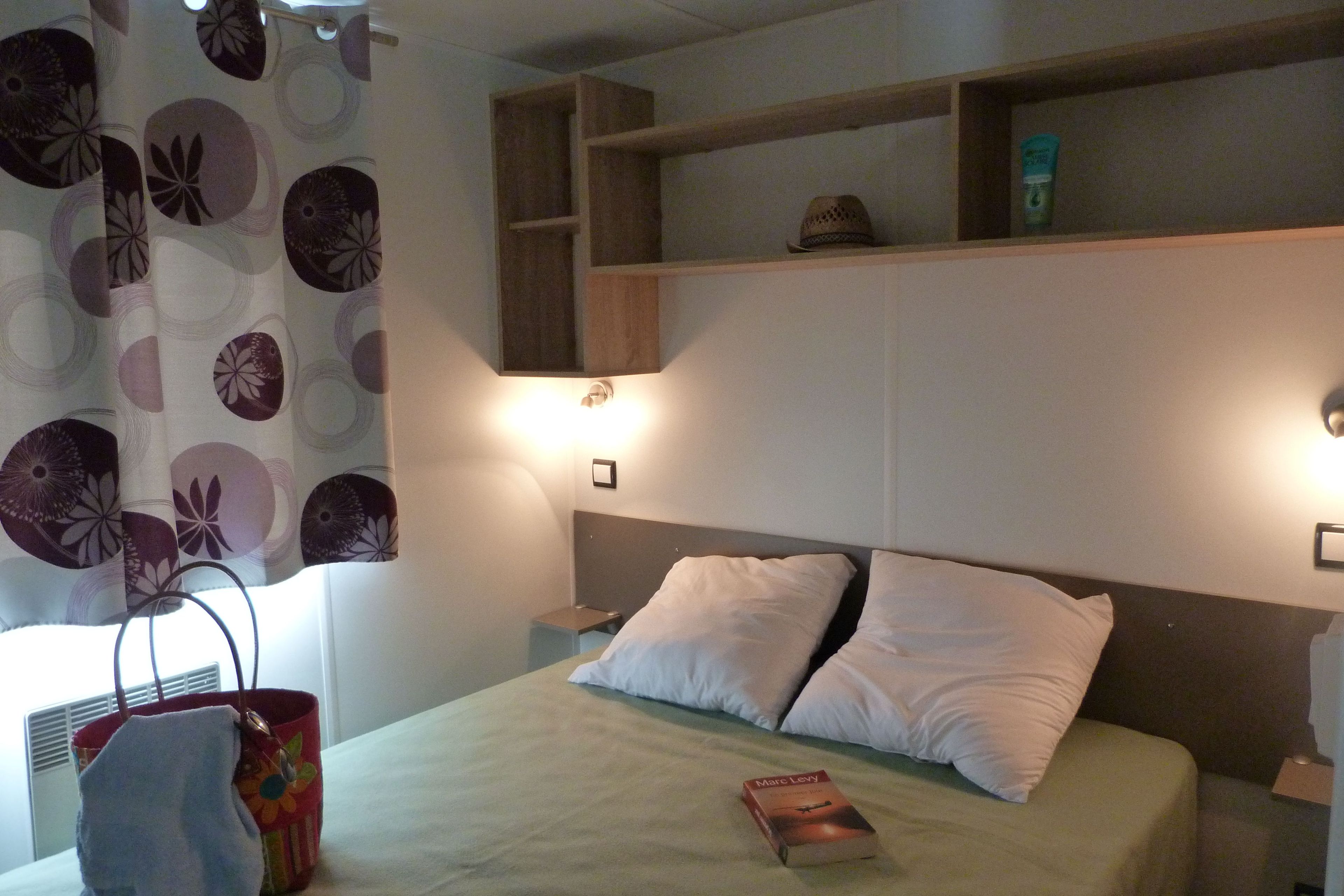 Une chambre d’hôtel avec un lit, une table de chevet, une étagère avec des articles de toilette et un rideau à motifs.