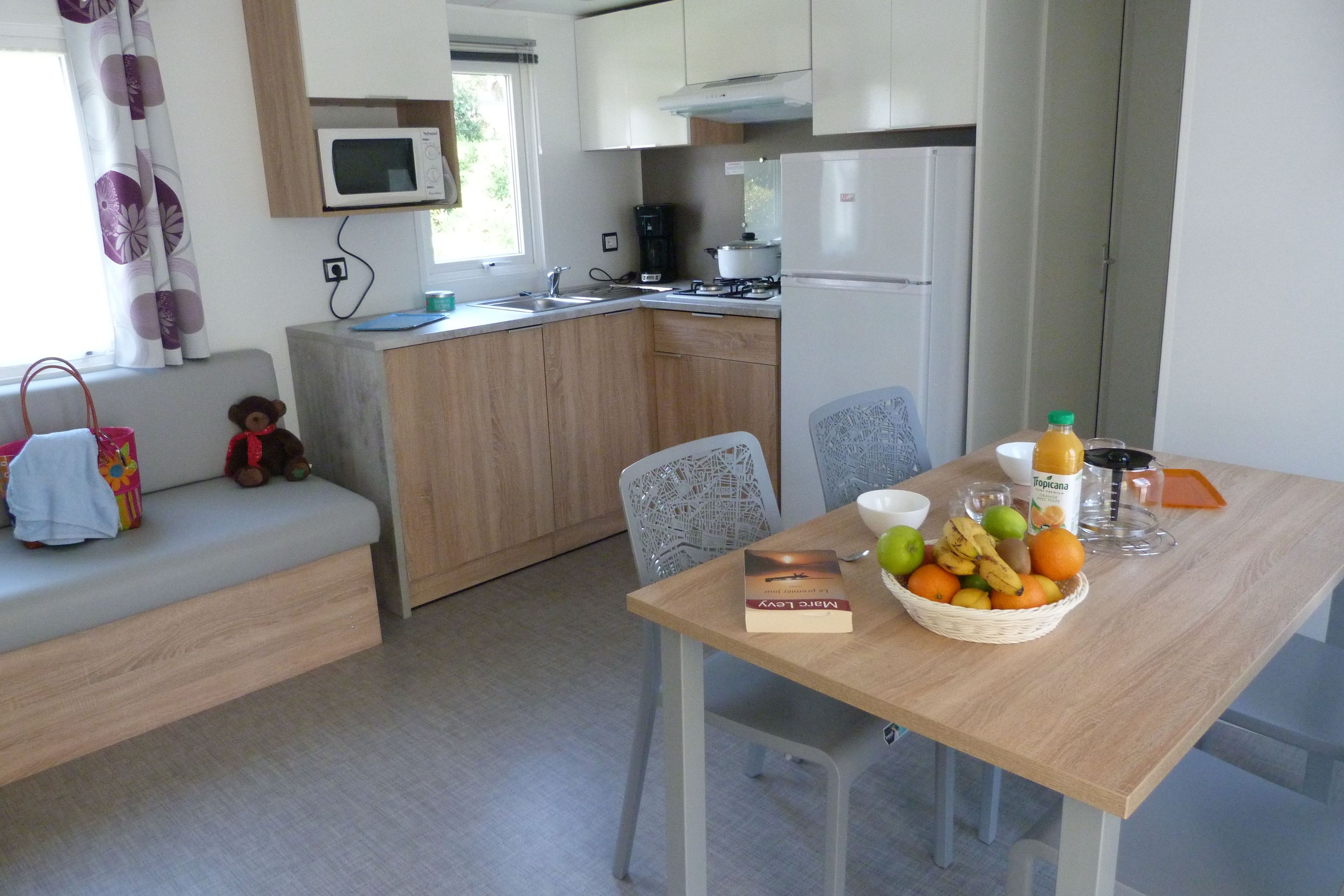 Cozinha compacta com micro-ondas, fogão e geladeira. Há uma mesa de jantar com uma cesta de frutas e um livro.