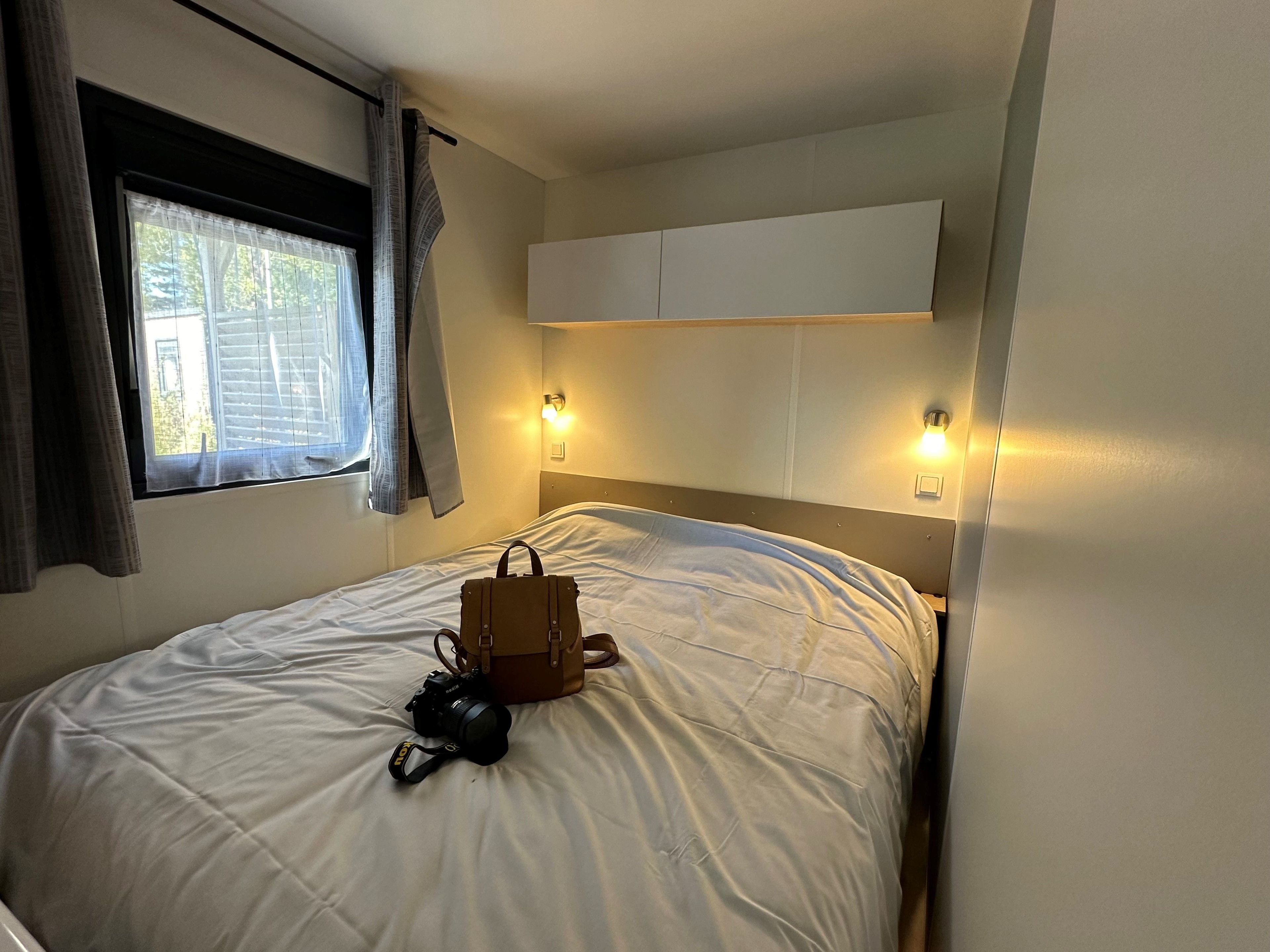 Ein Hotelzimmer mit einem Bett, einer Kamera und einer Handtasche auf dem Bett. An beiden Seiten des Bettes befinden sich zwei Wandleuchten.