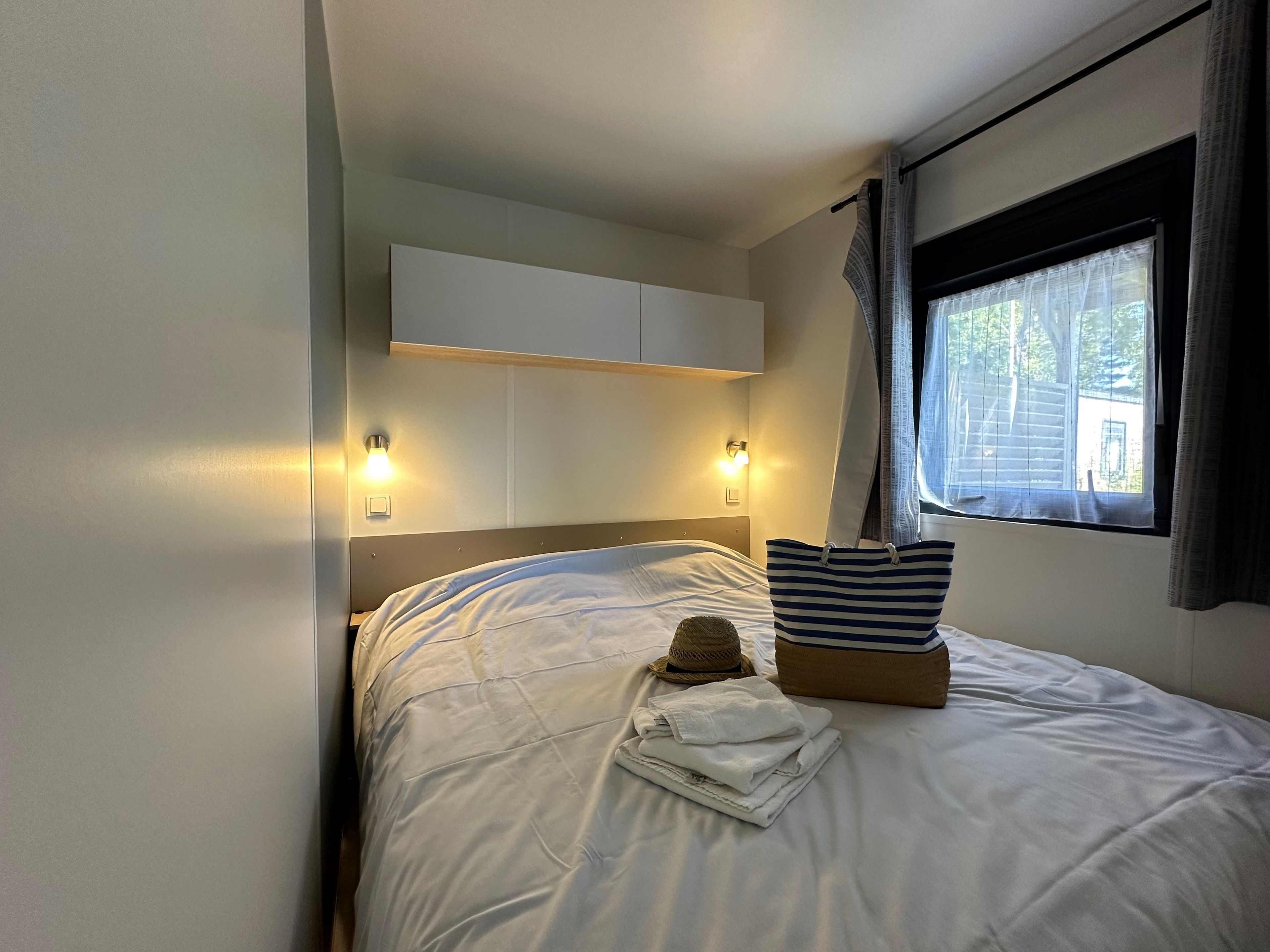 Een net opgemaakt bed met een gestreept kussen en een opgevouwen handdoek, een klein mandje en een strohoed. Aan weerszijden van het bed zijn twee wandlampen bevestigd.