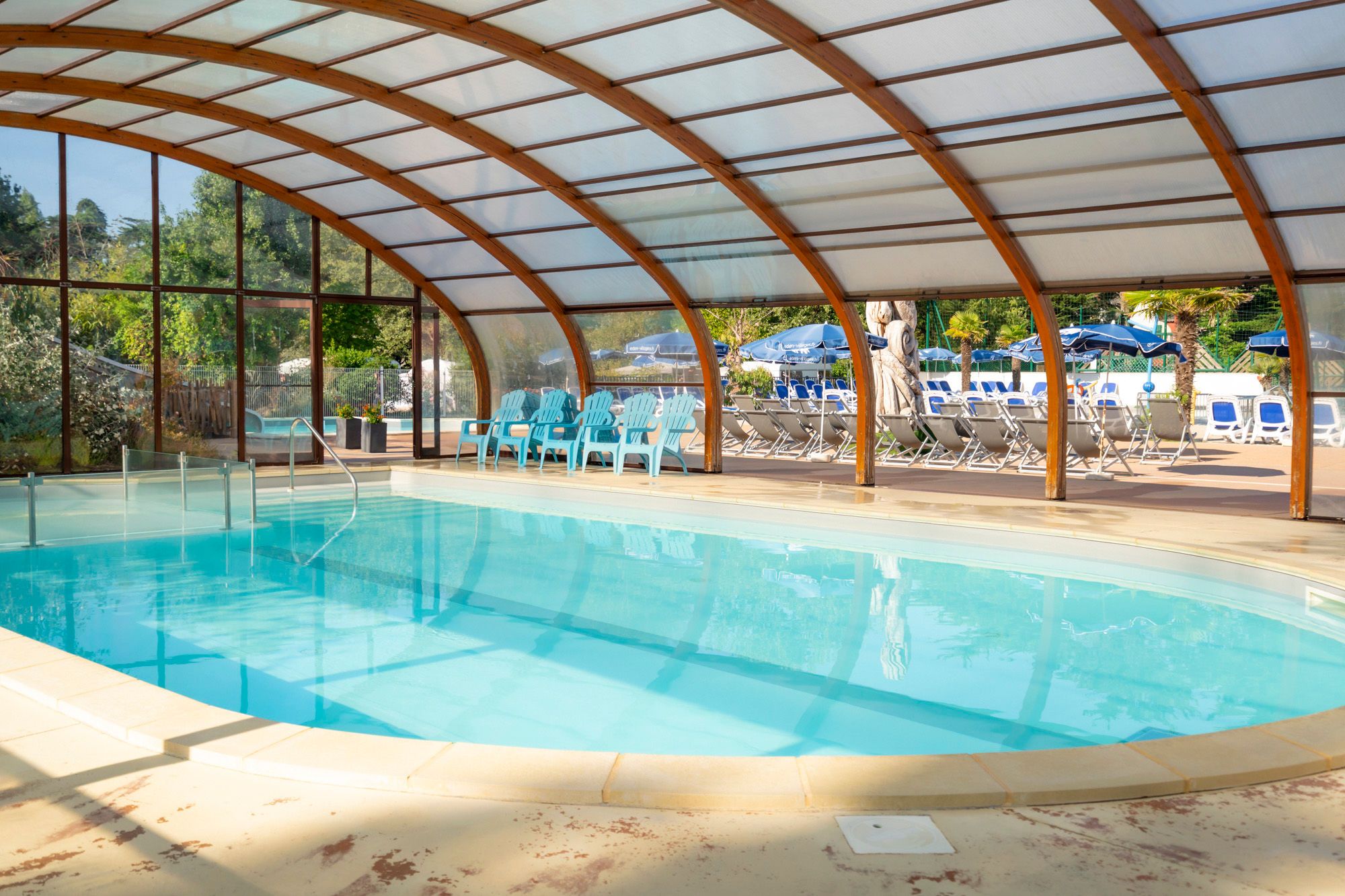 An indoor swimming pool with a glass ceiling, surrounded by lounge chairs and umbrellas.