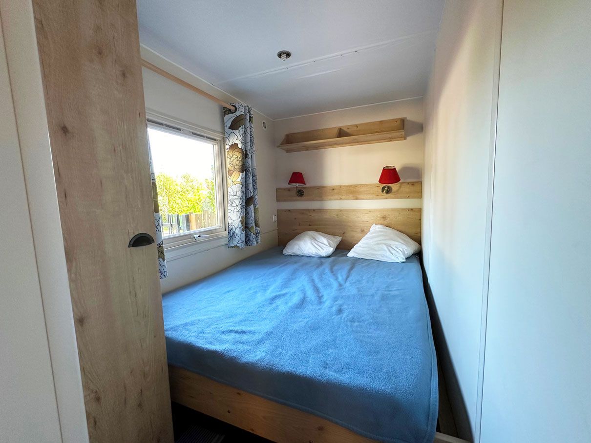 Un lit simple avec une couvre-lit bleue et deux oreillers, une fenêtre avec des rideaux et une tête de lit en bois.
