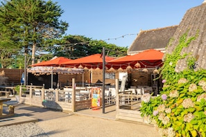 Een terras met tafels en stoelen, overdekt met rode parasols, en een stenen muur met beplanting.