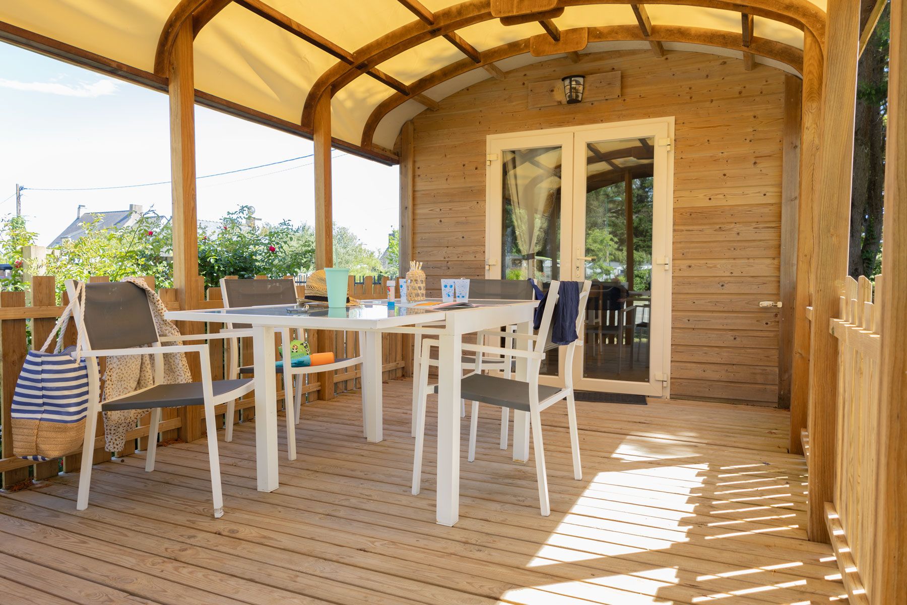 Une terrasse en bois avec une table et des chaises, un espace salon couvert et une porte donnant sur un espace extérieur.