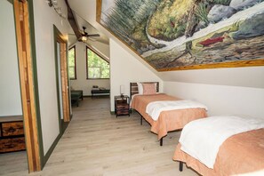 Une chambre avec deux lits, un ventilateur de plafond, un tableau représentant un paysage fluvial et une vue sur un jardin, visible à travers de grandes fenêtres.