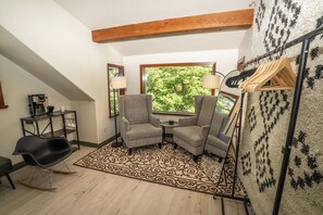 Een kamer met een hoge plafond, een kamerbreed tapijt met een patroon, twee fauteuils, een kleine tafel, een zwarte schommelstoel, een kapstok en een raam met uitzicht op bomen.