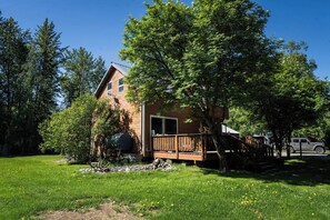 Une maison en bois à deux étages, dotée d’une terrasse, entourée d’arbres et bordée d’une pelouse.