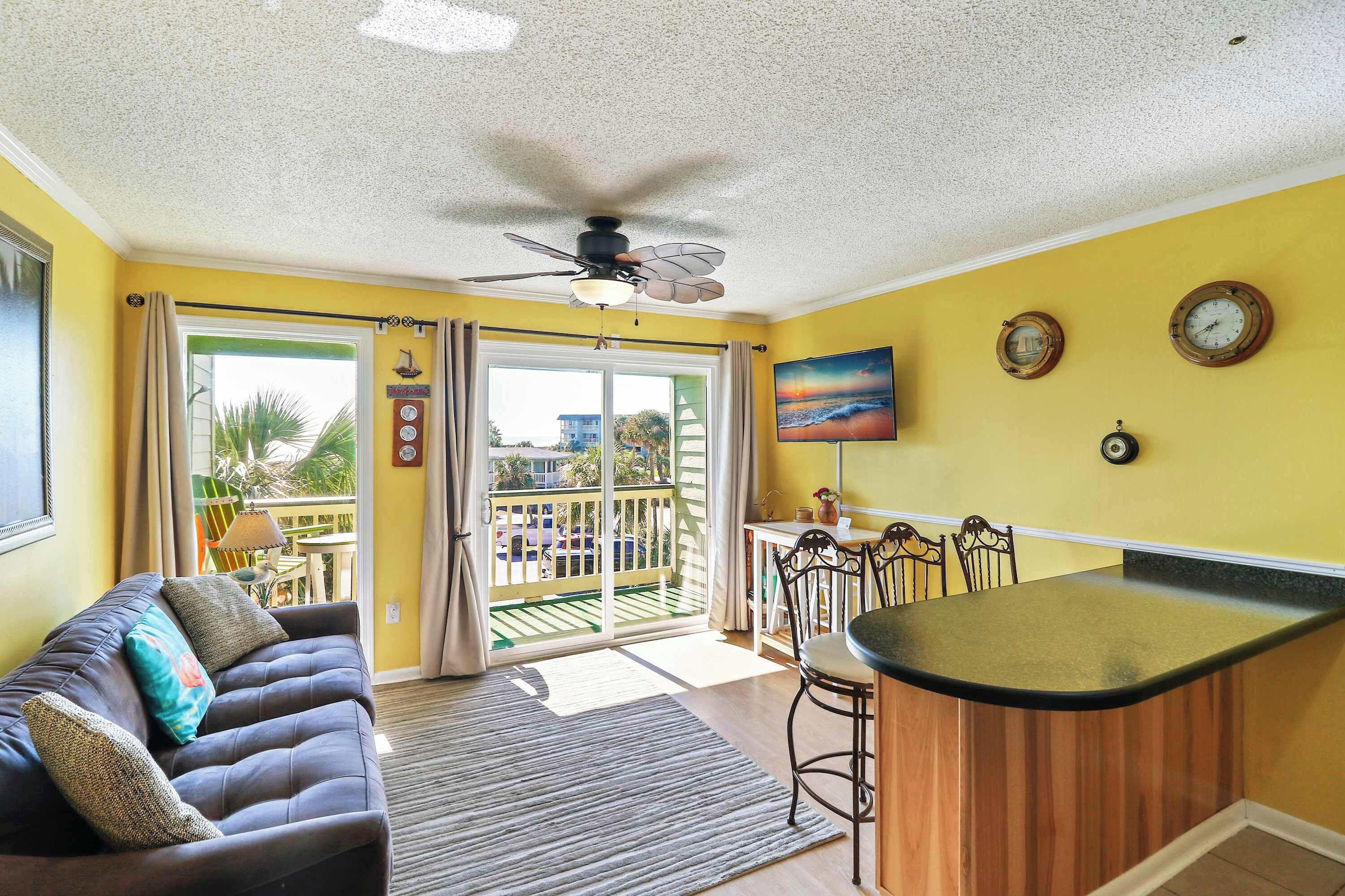 A room with a sofa, a dining area, a kitchen counter, and a view of the outdoors.