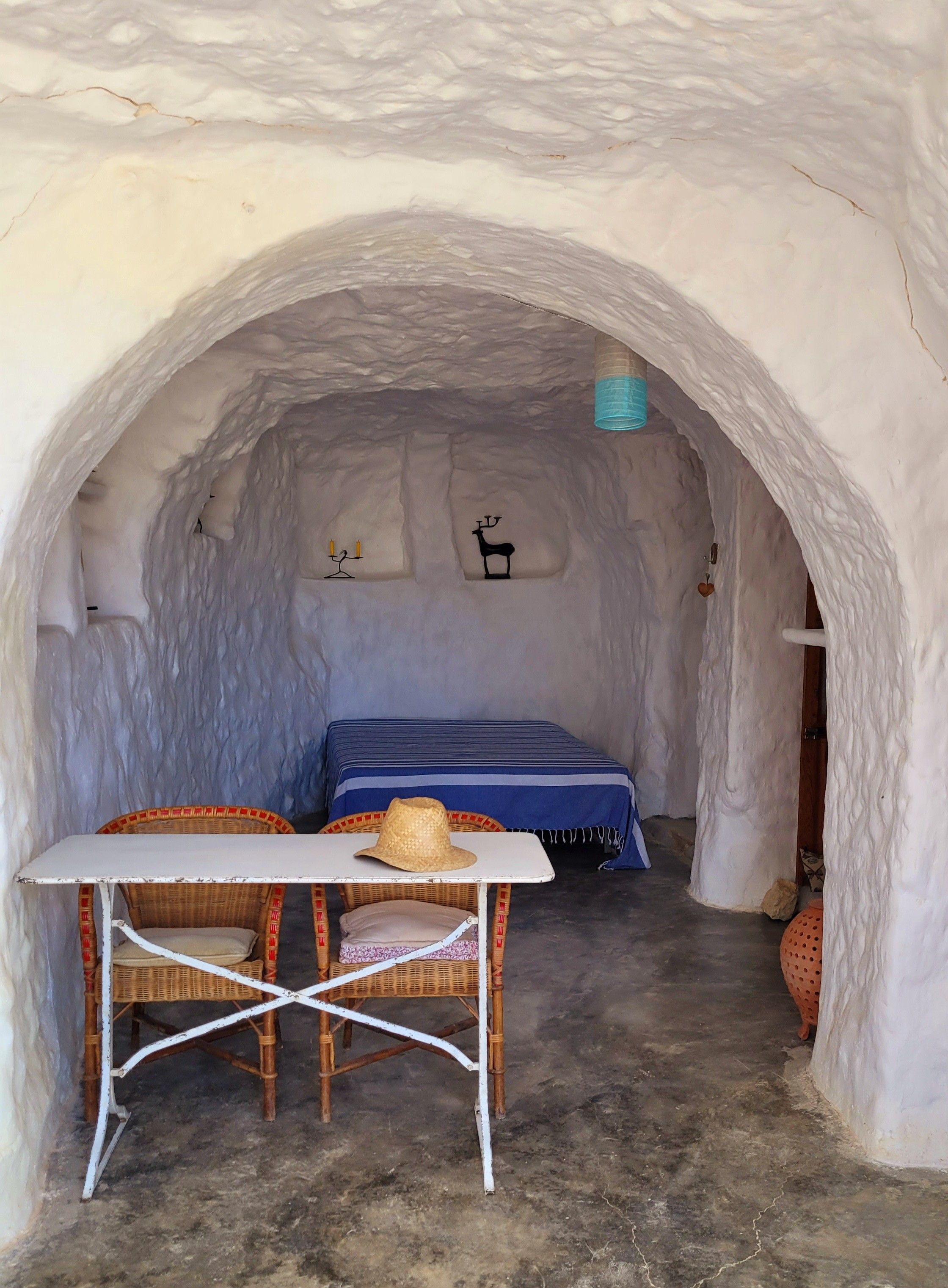 Una habitación acogedora con una cama, una mesa y una silla, todo dentro de una estructura parecida a una cueva, con una colcha azul y un sombrero de paja sobre la mesa.