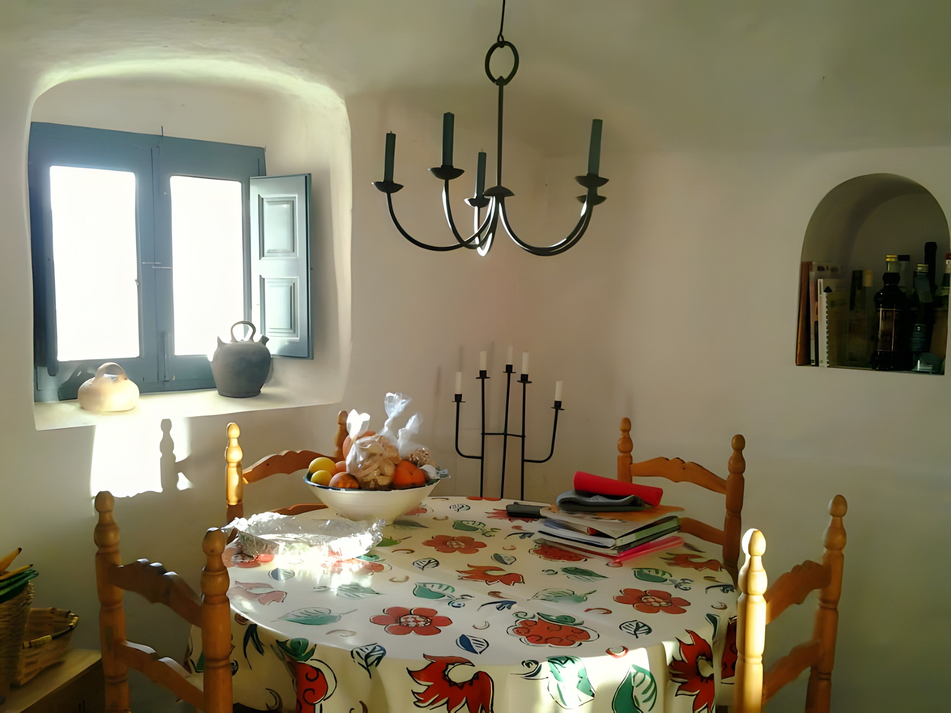Uma sala de jantar com uma mesa redonda, cadeiras, um candelabro, uma janela com persianas e uma estante na parede com garrafas.