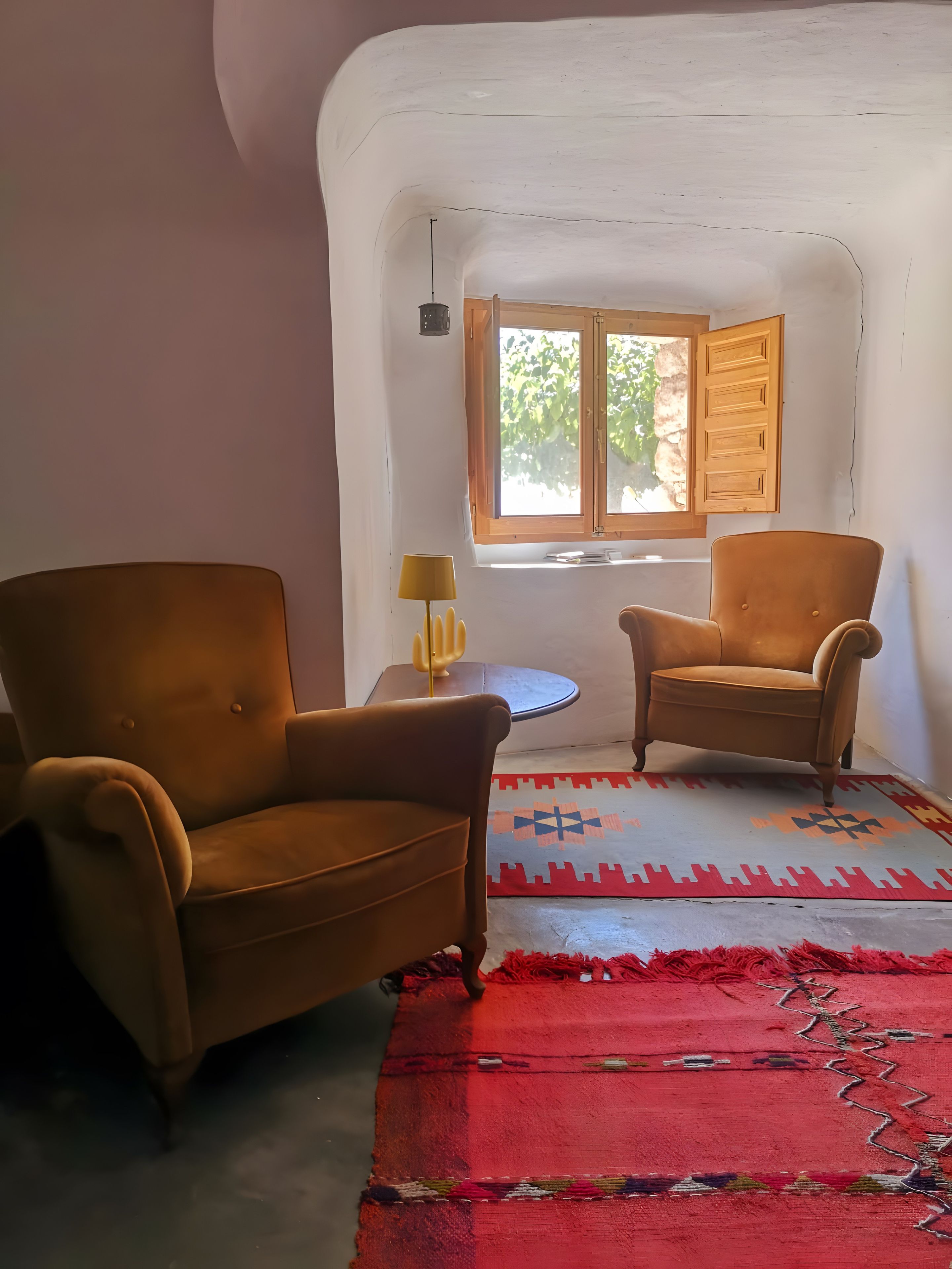 A room with two brown armchairs, a small table with a lamp, a patterned rug, and a window with wooden shutters.